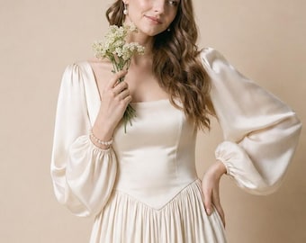 Puede incluir: Un vestido de satén color crema con escote cuadrado, mangas largas y voluminosas tipo obispo con puños fruncidos y un corpiño ajustado que se une a una falda plisada. La prenda se complementa con una pulsera y pendientes de perlas.