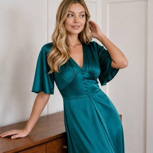 May include: A dark teal satin dress featuring a V-neckline, short flutter sleeves, and a row of matching fabric-covered buttons down the center of the bodice. The garment has a fitted waistline and a flowing skirt that reaches below the knees.