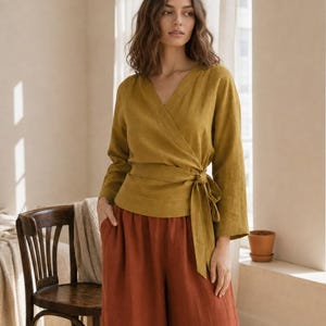May include: A mustard yellow long-sleeve wrap top featuring a V-neckline and a side tie closure, paired with loose-fitting rust-coloured wide-leg trousers. The top has a structured wrap design that secures at the waist with a fabric belt.