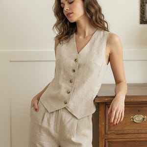Könnte beinhalten: Eine ärmellose Weste in Beige mit V-Ausschnitt und einer Knopfleiste mit fünf braunen Knöpfen auf der Vorderseite. Die Weste wird mit einer passenden Hose mit hoher Taille, Seitentaschen und einer Vorderfalte zu einem Set kombiniert.