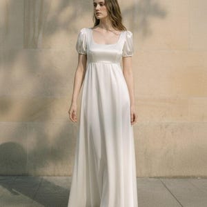 Puede incluir: Un vestido largo de satén color marfil con escote cuadrado, mangas cortas abullonadas y cintura imperio. La prenda presenta un acabado liso y brillante, con una falda fluida en forma de A que llega hasta el suelo, creando una silueta sencilla.
