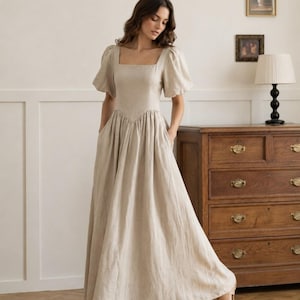 May include: A floor-length beige dress with a square neckline, short puff sleeves, and a fitted bodice that transitions into a gathered, full skirt. The dress features a V-shaped waistline seam and side pockets, crafted from a solid, light-coloured fabric.