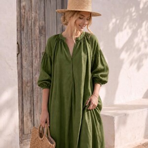 May include: A long olive green dress with a split neckline and voluminous three-quarter length sleeves with gathered cuffs. The dress is paired with a light brown straw hat and a matching woven straw handbag with circular handles.