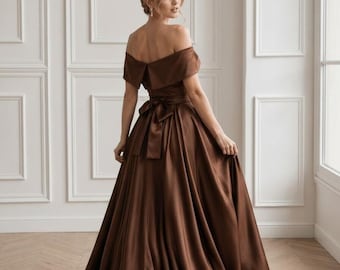Puede incluir: Un vestido largo de satén color marrón chocolate con escote de hombros descubiertos y detalle de cuello doblado. El vestido tiene un corpiño ajustado con un lazo atado en la cintura y una falda amplia y fluida que llega al suelo con acabado liso.