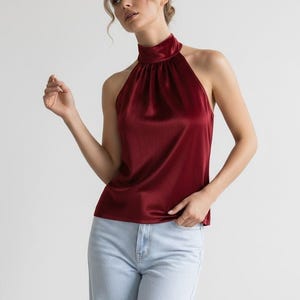 Puede incluir: Una blusa sin mangas de satén en color rojo oscuro con cuello alto y detalles de tela fruncida en el escote, combinada con pantalones vaqueros de color azul claro. La prenda tiene un acabado liso y reflectante con un ajuste ceñido al cuello.