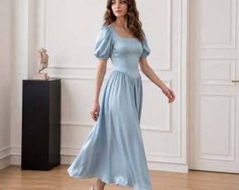 Puede incluir: Un vestido midi de color azul claro con escote cuadrado, mangas cortas abullonadas y un corpiño ajustado que da paso a una falda fluida en forma de A. La prenda está confeccionada en un tejido satinado liso con un brillo sutil, combinado con tacones beige.