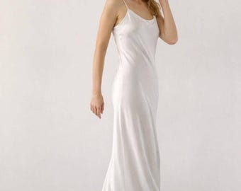 Puede incluir: Un vestido lencero blanco largo hasta el suelo con escote redondo y tirantes finos. La prenda está confeccionada con un tejido liso y brillante que cae a lo largo del cuerpo en una silueta fluida, extendiéndose hasta el suelo.