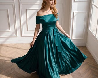 Puede incluir: Un vestido largo hasta el suelo, con hombros descubiertos, confeccionado en tela de satén de color verde esmeralda intenso. La prenda presenta un escote ancho y doblado, un corpiño ajustado y una falda amplia plisada con un cinturón de tela.