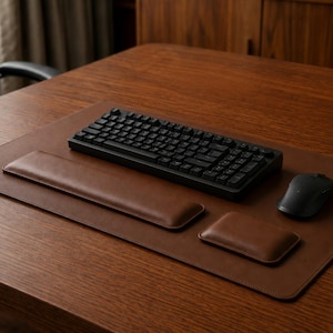 Op de afbeelding: Een bruine lederen bureauonderlegger met een bijpassende lange rechthoekige polssteun en een kleinere rechthoekige muispolssteun. Een zwart mechanisch toetsenbord en een zwarte draadloze computermuis zijn op het gladde bruine oppervlak geplaatst.