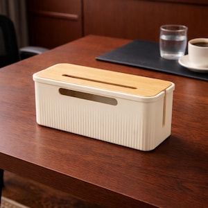 May include: A cream-colored cable management box with a bamboo lid sits on a dark wood surface. The box has a ribbed texture and a slot for cords. A glass of water and a cup of coffee are in the background.