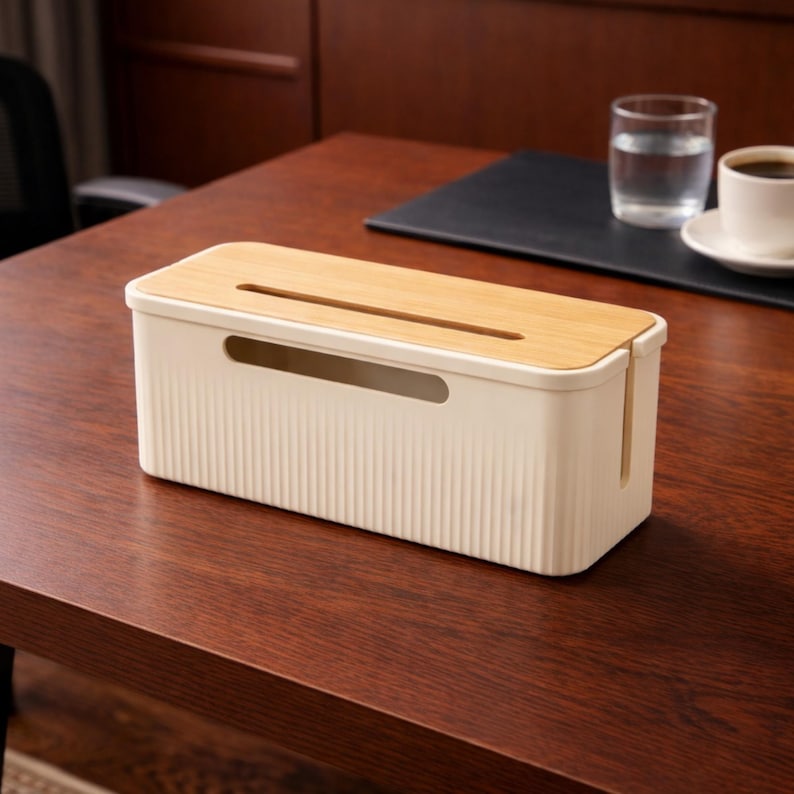 May include: A cream-colored cable management box with a bamboo lid sits on a dark wood surface. The box has a ribbed texture and a slot for cords. A glass of water and a cup of coffee are in the background.