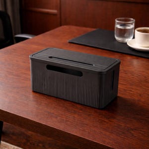 Puede incluir: Una caja negra para la gestión de cables con un diseño acanalado en los laterales y la parte superior, con una ranura para los cables. La caja está sobre un escritorio de madera, con un vaso de agua y una taza de café en el fondo. Este organizador ayuda a mantener los cables ordenados.