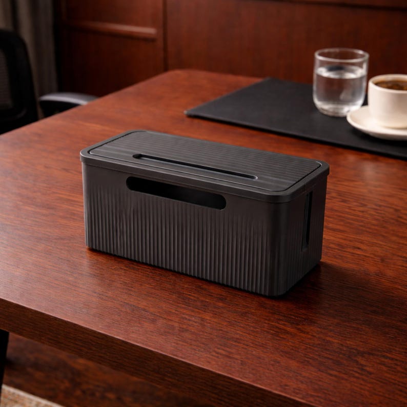 May include: A black cable management box with a ribbed design on the sides and top, featuring a slot for cords. The box is on a wooden desk, with a glass of water and a cup of coffee in the background. This organizer helps to keep cables tidy.