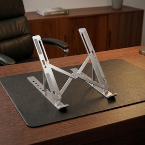 May include: A silver laptop stand with adjustable height settings. The stand is made of metal and has a foldable design. It is positioned on a black desk mat, with a wooden desk and office furniture in the background.
