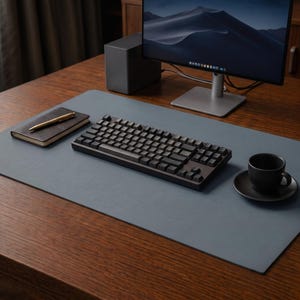 May include: A large, rectangular, slate-blue desk mat covering a wooden surface. Centered on the mat is a black mechanical keyboard. To the left sits a dark-covered notebook with a gold pen, and to the right is a black ceramic coffee cup and saucer.