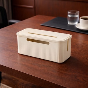 May include: A rectangular, off-white cable management box with a ribbed texture and a slotted top. The box has rounded corners and a smooth, hinged lid. It sits on a dark brown wooden surface, with a glass of water and a cup of coffee in the background.