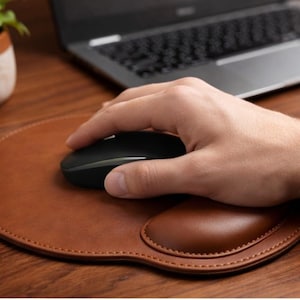 Può includere: Un mouse per computer wireless nero è appoggiato su un tappetino per mouse in pelle marrone con un poggiapolsi integrato. Il tappetino è su una scrivania di legno, con un computer portatile sullo sfondo.