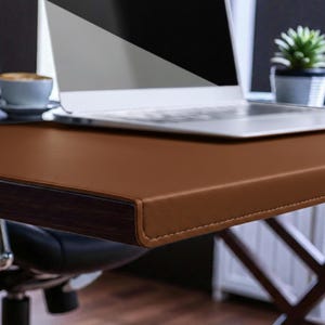 May include: A brown leather desk with a dark wood trim and a laptop. The desk has a stitched edge and is set up in an office setting. A small potted plant and a cup of coffee are also visible.
