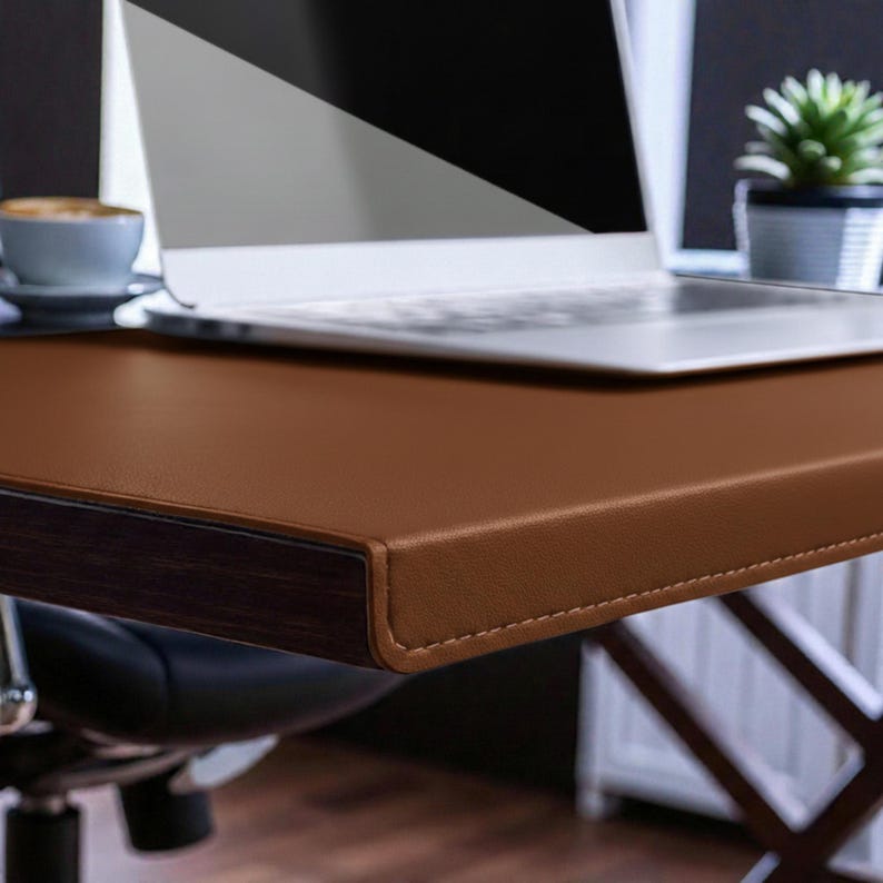 May include: A brown leather desk with a dark wood trim and a laptop. The desk has a stitched edge and is set up in an office setting. A small potted plant and a cup of coffee are also visible.