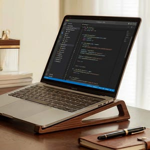 Può includere: Un computer portatile argentato che mostra righe di codice Python sullo schermo, appoggiato su un supporto in legno marrone scuro. Una penna stilografica nera è appoggiata su un taccuino con copertina in pelle marrone in primo piano.