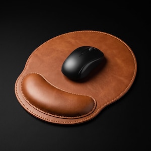 May include: A tan leather mouse pad with a built-in wrist rest and visible stitching along the edges. A black wireless computer mouse is positioned on the smooth surface of the pad. The design features a rounded shape with a curved support section.