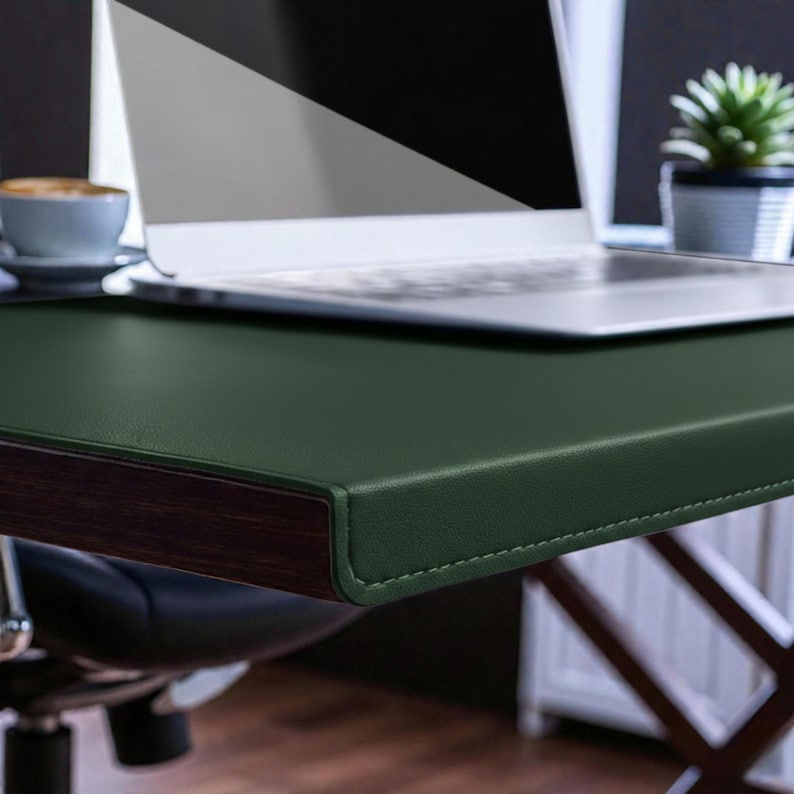 May include: A dark green desk with a laptop, a cup of coffee, and a small potted plant. The desk has a dark wood trim and a stitched leather surface. The laptop is open, and the background is blurred.