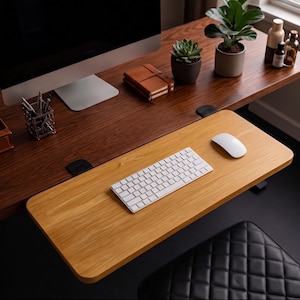 Op de afbeelding: Een bureauverlenger van licht hout bevestigd aan een donkerhouten bureau. De verlenger bevat een wit draadloos toetsenbord en een witte computermuis. Het hoofdbureau bevat een computermonitor, een metalen pennenhouder, een bruin notitieboek en twee planten.
