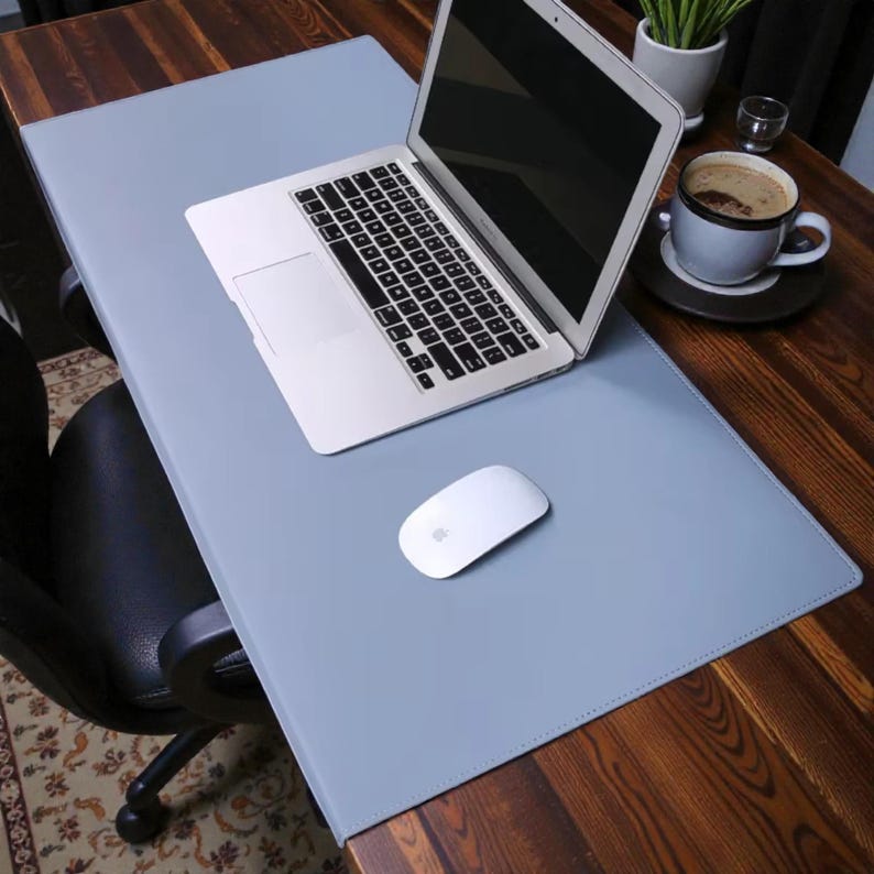 May include: A light gray desk mat with a laptop and a white mouse. The laptop is open, displaying a black screen. A cup of coffee sits on a saucer to the right. The desk is made of wood.