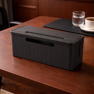 May include: A black cable management box with a ridged design on the sides and top. The box has a rectangular shape with a lid and a handle cutout on the side. It sits on a wooden desk.