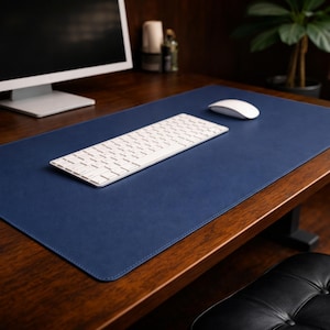 Puede incluir: Una alfombrilla de escritorio azul marino con un teclado y un ratón blancos sobre un escritorio de madera. La alfombrilla es rectangular y cubre una gran parte de la superficie del escritorio. El teclado es de tamaño estándar con teclas blancas. Un monitor de ordenador es visible en el fondo.