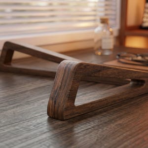 May include: A dark brown wooden laptop stand featuring a triangular frame design with visible wood grain patterns. The stand consists of two separate angled supports that provide a raised platform for a device, showcasing a smooth, polished finish.