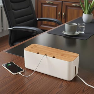 May include: A white cable management box with a light wood grain lid sits on a wooden desk. A smartphone is charging next to the box. The desk also has a black desk pad, a cup, and a glass of water.