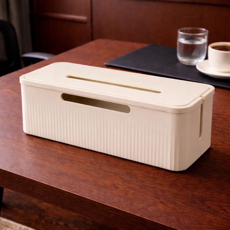 May include: A cream-colored cable management box with a ribbed texture. The rectangular box has a slot on top and a curved opening on the side. It sits on a dark brown wooden surface, with a glass of water and a cup of coffee in the background.