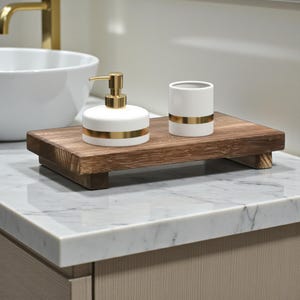 Könnte beinhalten: Ein hölzernes Badezimmer-Tablett mit einem weißen Seifenspender und einem passenden Becher, beide mit goldenen Bändern. Das Tablett ist aus dunklem Holz gefertigt und steht auf einer Marmorarbeitsplatte.