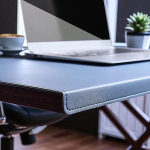 May include: A close-up of a modern desk with a gray leather-like surface and dark wood trim. A laptop, a small potted plant, and a cup of coffee are on the desk. The desk is supported by a dark wooden frame.