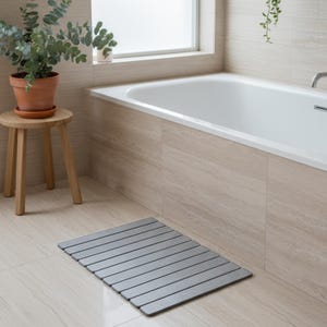Può includere: Un tappetino da bagno rettangolare grigio con linee orizzontali è posizionato su un pavimento piastrellato in un bagno. Il tappetino è di fronte a una vasca da bagno bianca. Un piccolo sgabello di legno con una pianta in vaso è a sinistra.