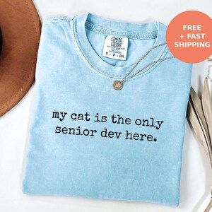 May include: A light blue short-sleeve t-shirt with a crew neckline. The shirt features the black printed text "my cat is the only senior dev here." A silver-toned necklace with a small circular pendant rests on the center of the chest area.