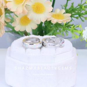 May include: A pair of silver hoop earrings with white opal inlays. The earrings are displayed on a white, textured surface with the text "SHAZMABEAUTYGEMS" and "Handcrafted Jeweller" in the background. The background also features flowers.
