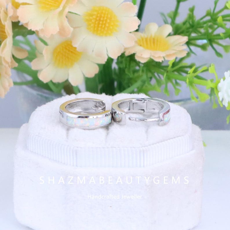 May include: A pair of silver hoop earrings with white opal inlays. The earrings are displayed on a white, textured surface with the text "SHAZMABEAUTYGEMS" and "Handcrafted Jeweller" in the background. The background also features flowers.