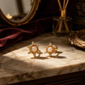 May include: Two gold-colored stud earrings. Each earring features a central, round, off-white stone surrounded by small, clear, sparkling stones. The earrings have a starburst design and are displayed on a marble surface.
