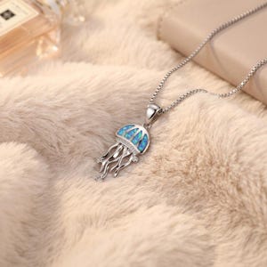 May include: A silver necklace featuring a jellyfish pendant. The pendant has a blue and white opal design with clear accent stones. The jellyfish has flowing tentacles and is suspended from a delicate chain. The necklace is displayed on a soft, textured surface.