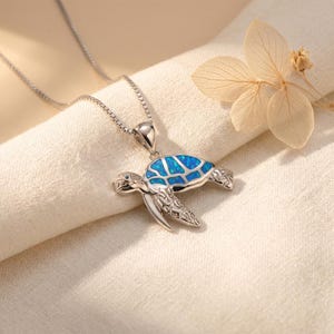 May include: A silver-colored turtle pendant necklace with a blue opal inlay. The turtle has detailed flippers and head, and the pendant hangs from a delicate chain. The necklace is displayed on a cream-colored fabric.