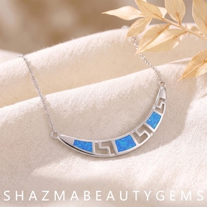 May include: A silver-toned crescent-shaped necklace pendant featuring a repeating geometric Greek key pattern interspersed with inlaid iridescent blue opal-like segments, suspended from a delicate matching silver-toned metal chain.