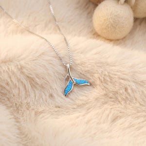 Può includere: Una collana argentata con un ciondolo a forma di coda di balena. La coda è riempita di materiale blu simile all'opale. La collana è esposta su una superficie soffice e chiara.