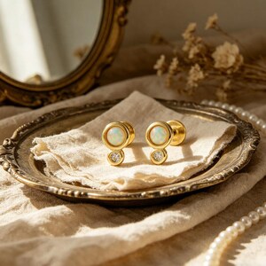 Può includere: Un paio di dilatatori per orecchie color oro con centri in opale e una piccola gemma trasparente sotto. Sono esposti su un piatto decorativo in stile antico con un panno color crema.