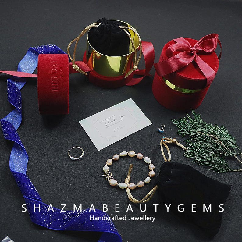 May include: An assortment of jewelry and gift packaging on a black surface. Items include a pearl bracelet, a ring, velvet boxes, a gold-colored container, a thank you card, and a blue ribbon with glitter.