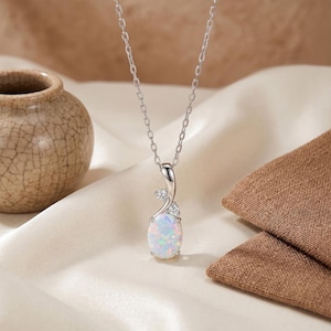 May include: A silver necklace with an oval opal pendant. The pendant is white with hints of pink and blue, and is accented with small clear crystals. The necklace is displayed on a cream-colored fabric.