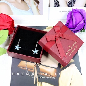 May include: A pair of starfish earrings with blue and silver accents displayed in an open red gift box. The box is next to a closed red gift box with a bow and the text "BEST WISHES FOR YOU, Especially for you."