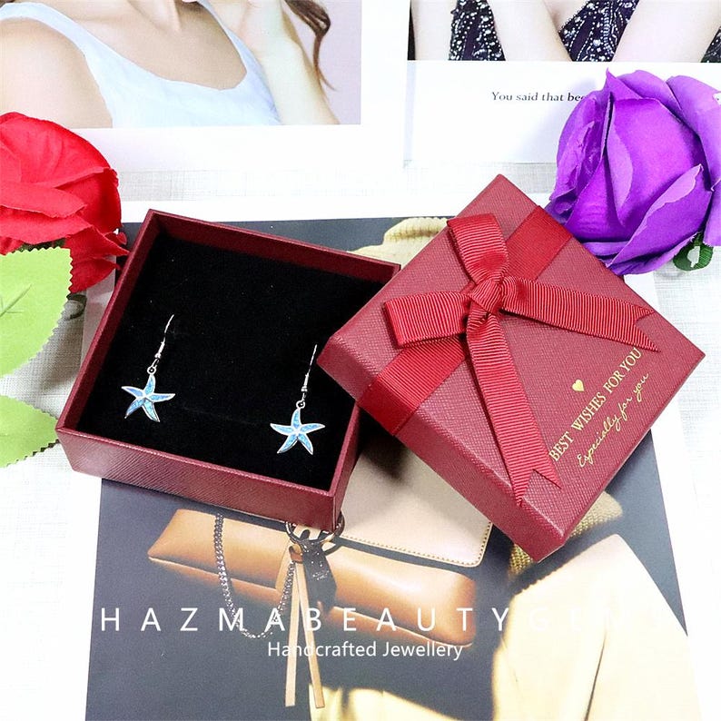 May include: A pair of starfish earrings with blue and silver accents displayed in an open red gift box. The box is next to a closed red gift box with a bow and the text "BEST WISHES FOR YOU, Especially for you."