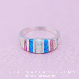 May include: A silver ring with a band of rectangular opal stones in shades of blue, white, and pink. The ring is set against a soft pink background. The text "SHAZMABEAUTYGEMS Handcrafted Jewellery" is at the bottom.